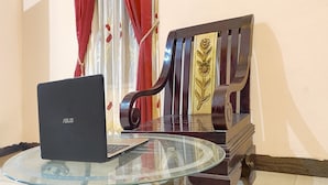 Una computadora portátil sobre una mesa de vidrio, una silla de madera con tapicería floral y cortinas rojas en una habitación con pared lisa.