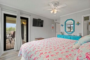 Un dormitorio con cama, ventilador de techo, televisor, cómoda y puerta corrediza a un balcón.