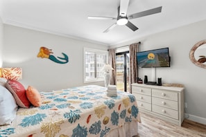 Ein Schlafzimmer mit einem Bett, einem Deckenventilator, einem Fernseher, einer Kommode und einem Fenster mit Jalousien.