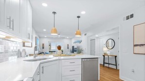 Une cuisine moderne avec des meubles blancs, un îlot central doté d’un lave-vaisselle, un éclairage suspendu et une vue sur la salle à manger.