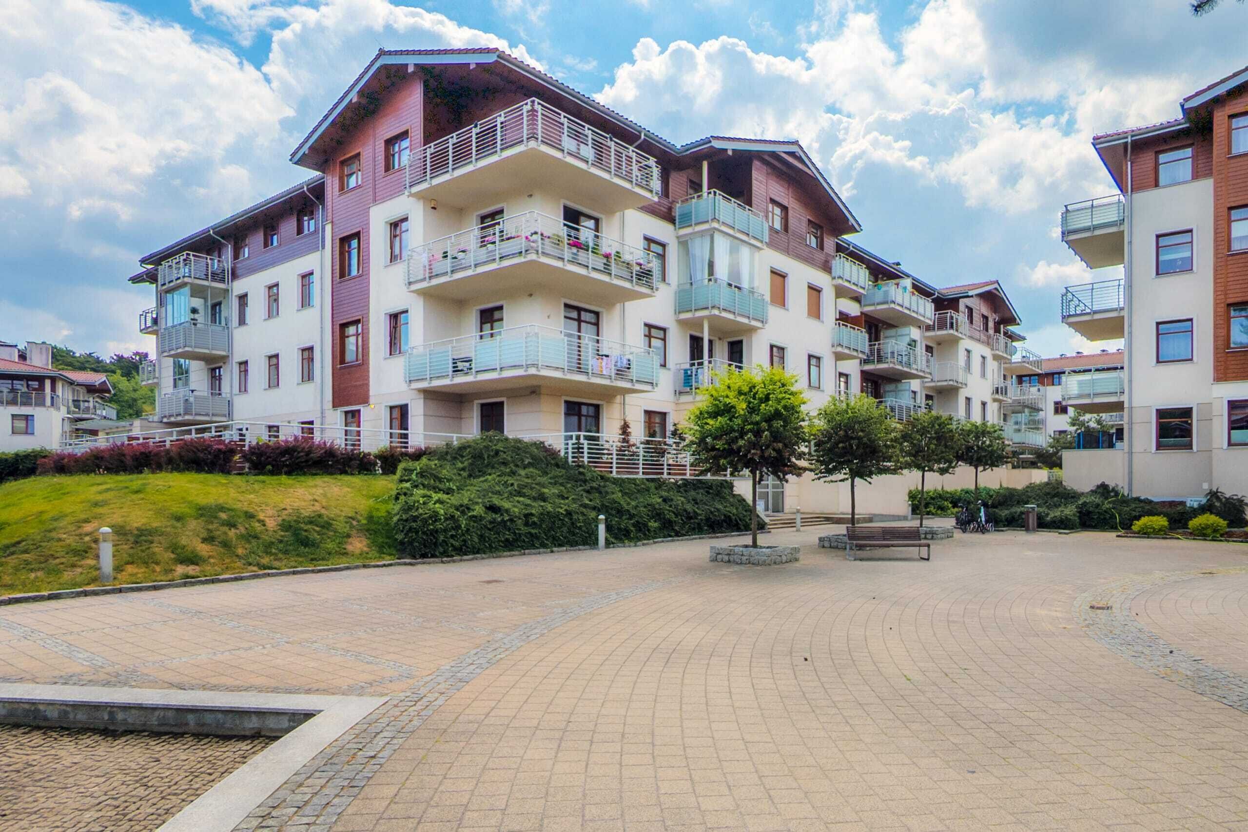 Un immeuble résidentiel à plusieurs étages, doté de balcons, d’une cour pavée et entouré de verdure.