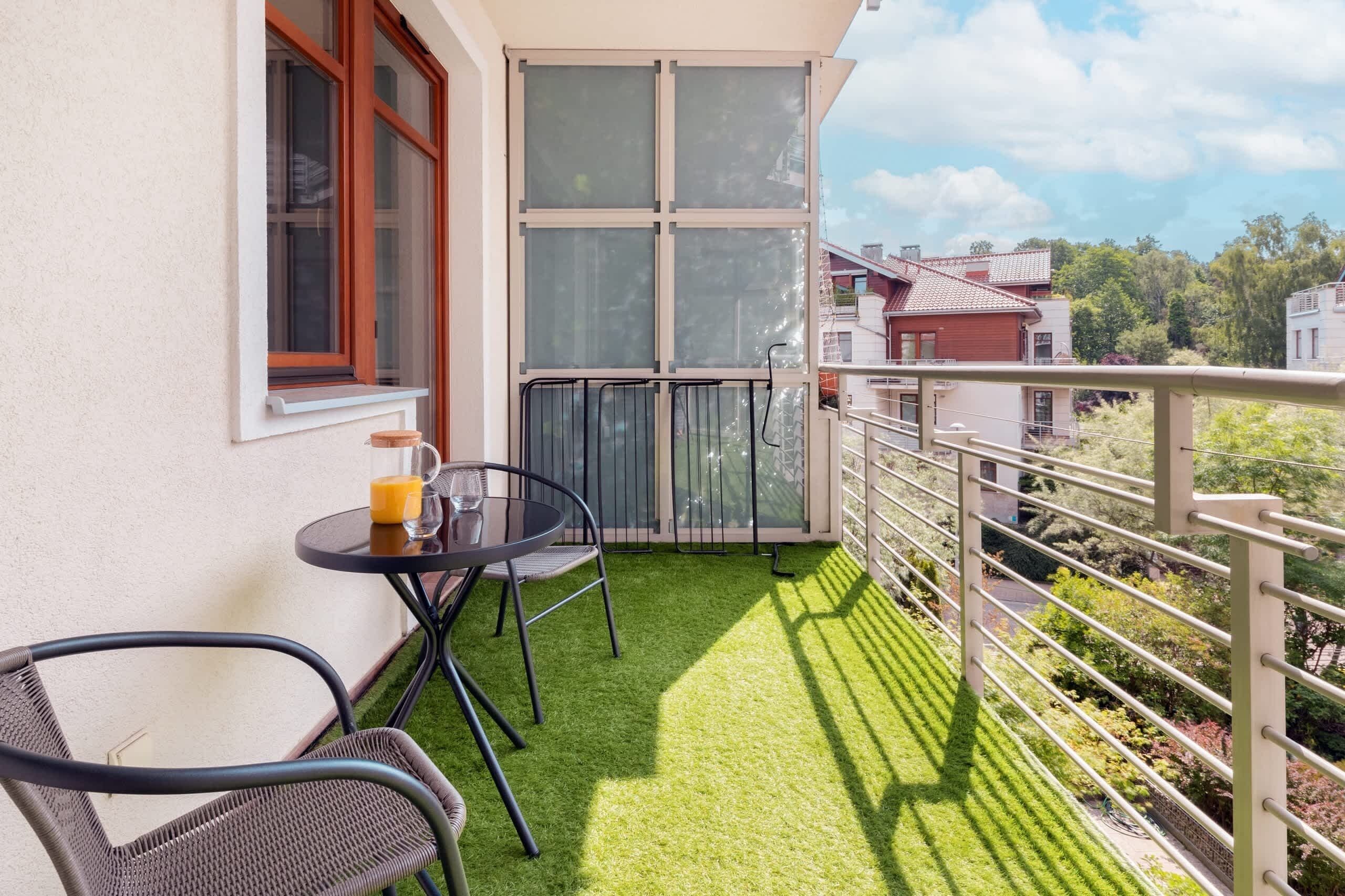 Ein Balkon mit Kunstgrasanlage, einem Tisch mit Stühlen, einem Glas Orangensaft und einem Fenster mit Jalousien.