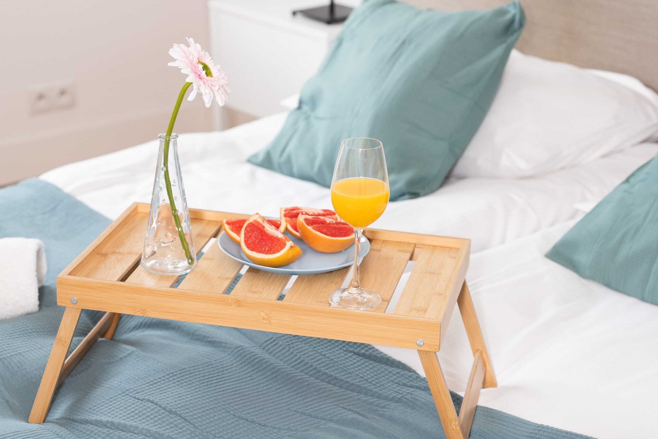 A wooden tray with a glass of orange juice, a plate with grapefruit halves, and a vase with a flower on a bed with white and teal pillows.