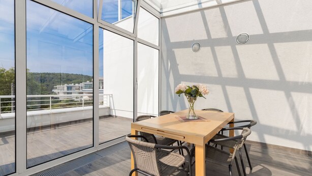 Un espace de salle à manger moderne, avec une table et des chaises, de grandes fenêtres et une vue sur un immeuble et des espaces verts.