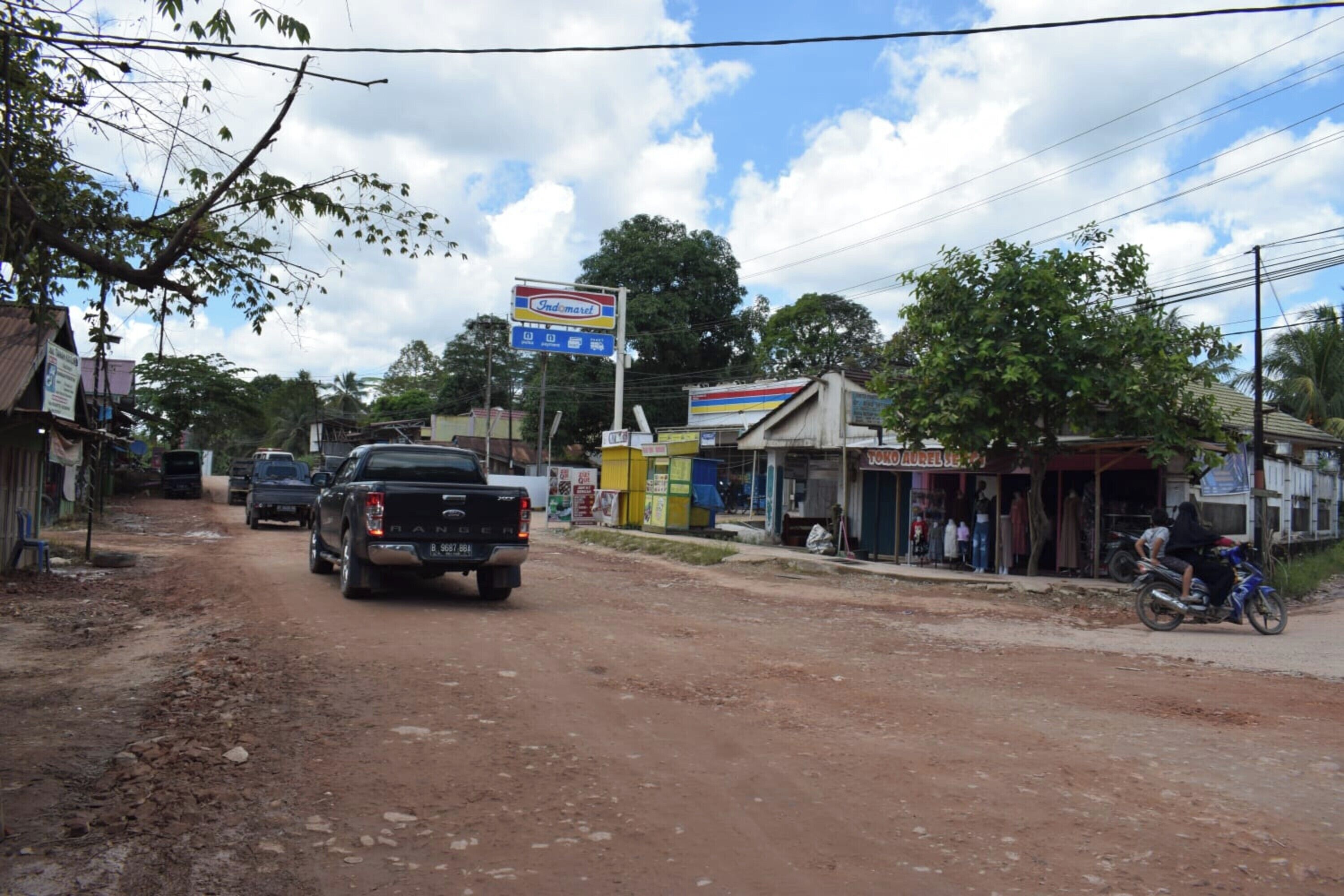 Un camino de tierra con una camioneta pickup y una motocicleta. Hay pequeñas tiendas y edificios a un lado del camino.