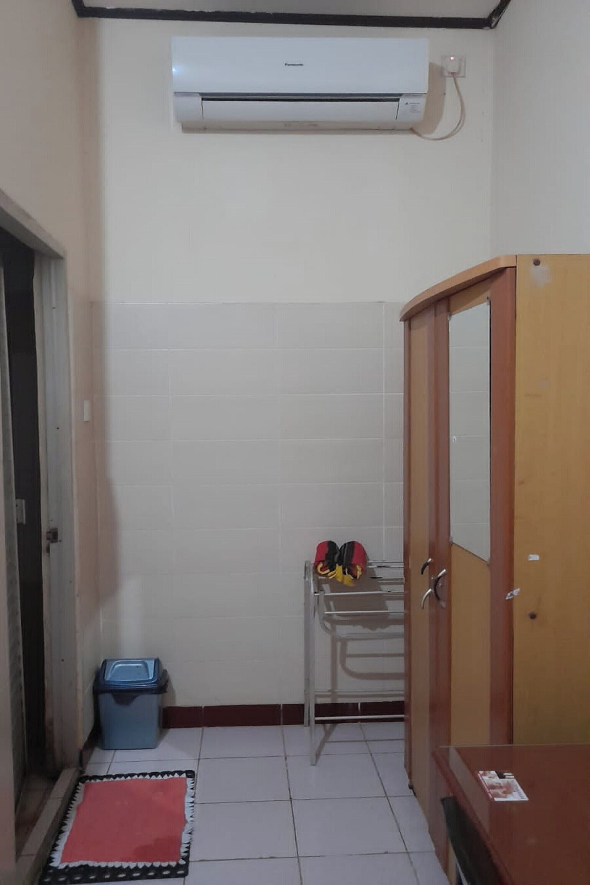 Una habitación pequeña con suelo de baldosas, un armario de madera, una estantería metálica con bolsos, un cubo de basura azul y una alfombra roja.