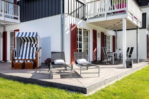 En toetagers bygning med balkon, udendørs siddeområde med liggestole og en stribet markise, samt et lille bord med stole.