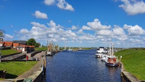 A canal with boats docked on both sides, surrounded by grassy banks and buildings with red roofs. - 1 - 4 Personen EG, Haustiere Willkommen Terrassenausrichtung Süd und West 2 Schlafzimmer, 1 Badezimmer mit Whirlpool und Dusche (Greetsiel)