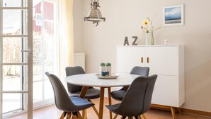 A dining area with a round table, four chairs, a cabinet with 'AZ' letters, a hanging light fixture, and a window with curtains. - 1 - 4 Personen EG, Haustiere Willkommen Terrassenausrichtung Süd und West 2 Schlafzimmer, 1 Badezimmer mit Whirlpool und Dusche (Greetsiel)