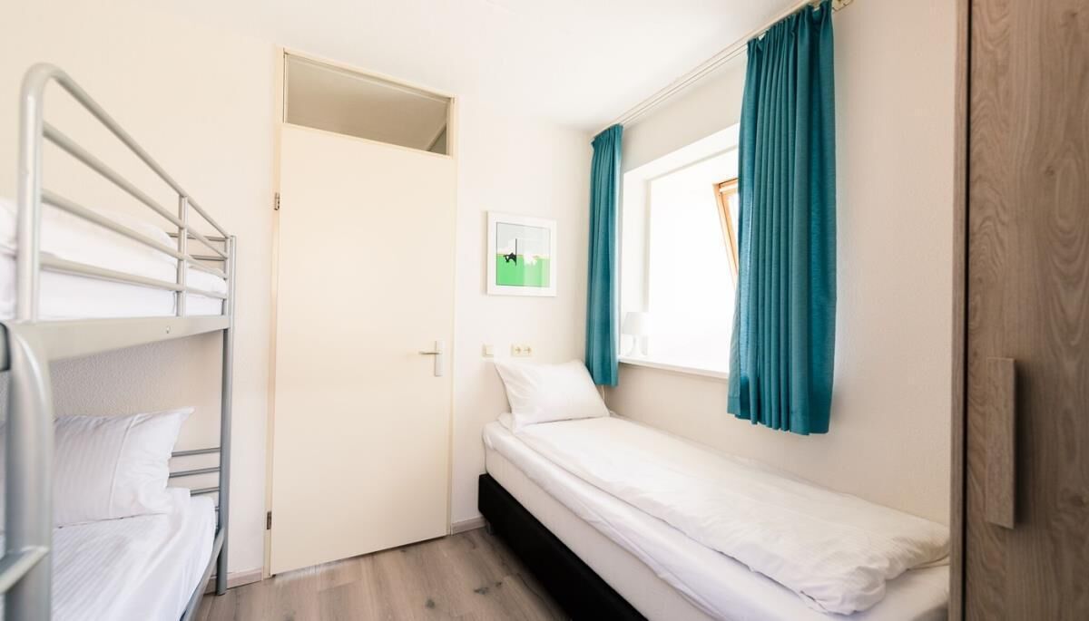 Ein kleines Zimmer mit einem Einzelbett und einem Bunkelbett links, türkisblauen Vorhängen und einem Fenster mit Blick.