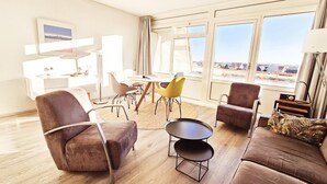En moderne stue med sofa, lænestole, et sofabord og et spiseområde med stole og bord. Store vinduer giver udsigt til det fri.