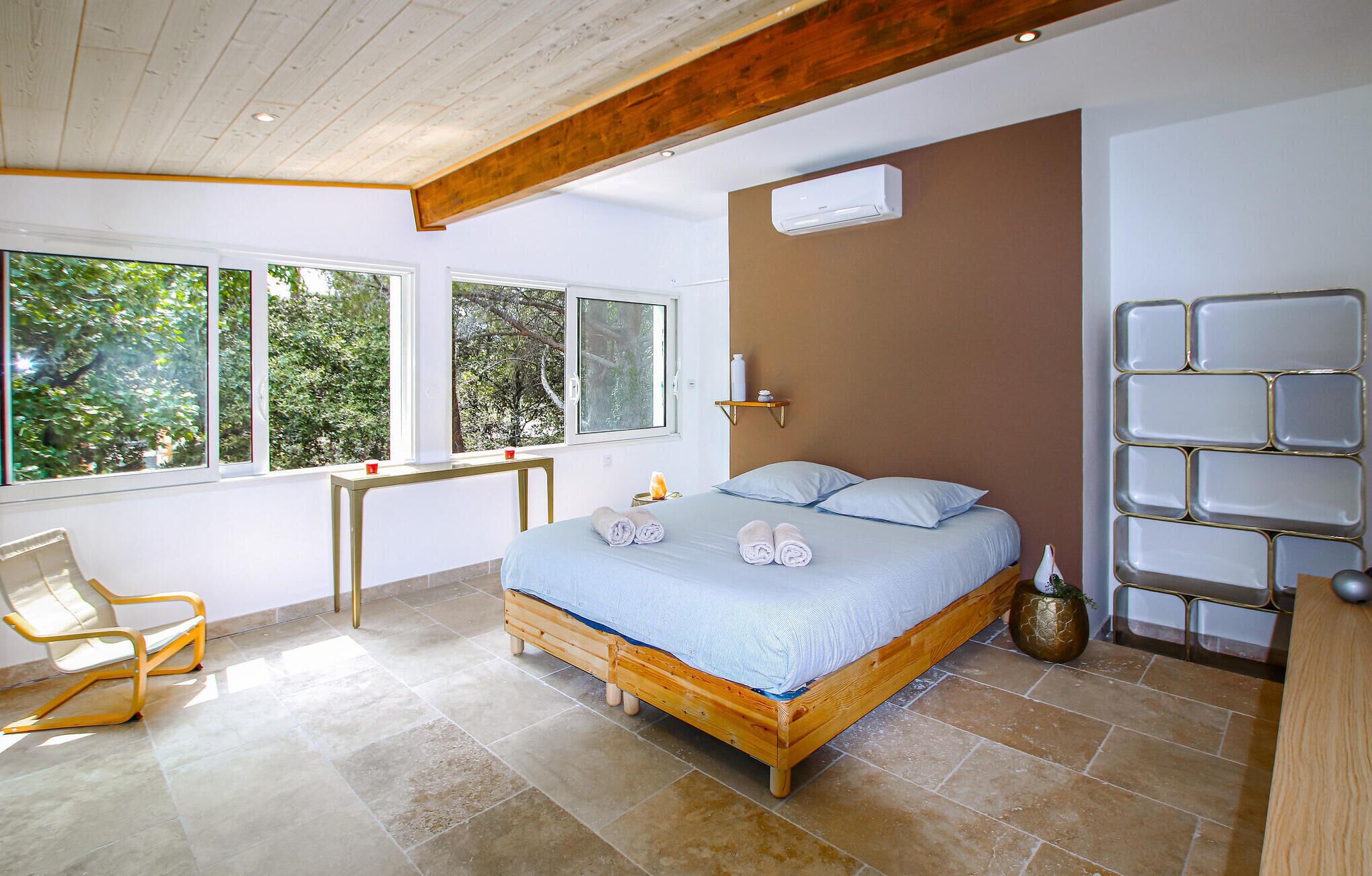 A bedroom with a wooden bed, a bedside table, a chair, and a window with a view of trees.