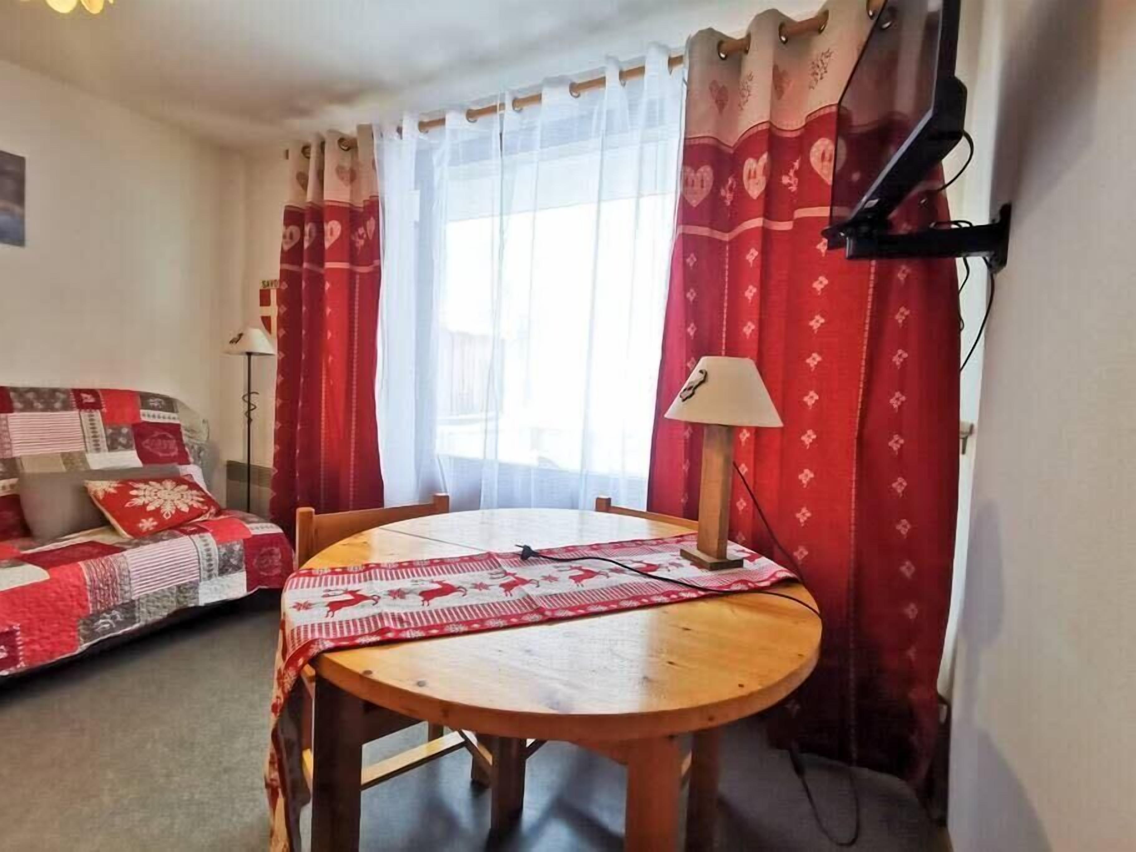 Una habitación acogedora con una mesa redonda de madera, un sofá con una manta de cuadros rojos y blancos, y una ventana con cortinas translúcidas.