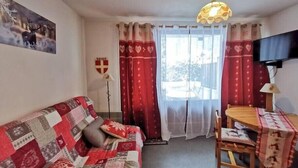 Een gezellige woonkamer met een gestreept bankje, een eettafel met een rode tafelkleed en een aan de muur bevestigde televisie.