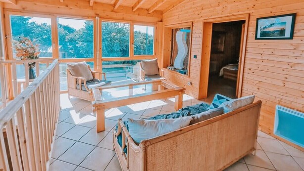 Interior de una cabaña de madera con balcón, muebles de mimbre y ventanas amplias.