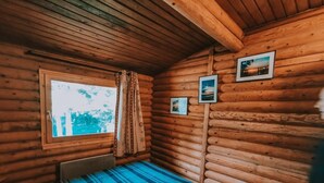 Una habitación tipo cabaña de madera con una cama, un ventanal con cortinas y tres cuadros en la pared.