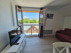 Una habitación con un ventanal grande, un televisor sobre un mueble bajo, un sofá rojo y una silla blanca.