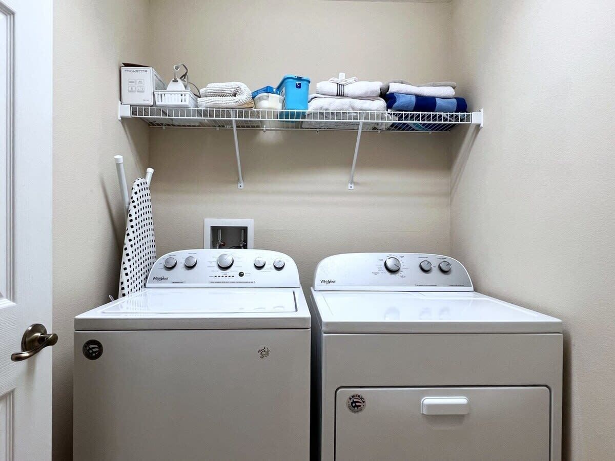 A laundry room with two Whirlpool washers and dryers, a shelf with laundry supplies, and a drying rack.