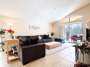 A modern living room with a dark grey sofa, a wooden coffee table, and a side table with books. There is a large window providing natural light and a view of the outdoors.