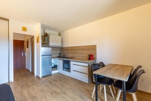 Een kleine keuken met een koelkast, fornuis en wastafel, een eettafel met stoelen en een houten accentmuur.