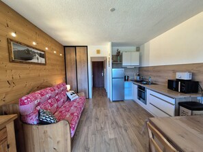 Een compact leefruimte met een houten wand, een bank, een keukenhoek met een koelkast, magnetron en broodrooster, en een houten eettafel.