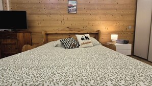 Ein Schlafzimmer mit Holz-Kopfteil, einem Bett mit gemusterten Bettbezügen, zwei Kissen, einem Nachttisch mit Lampe und einem Fernseher auf einer Holzkommode.