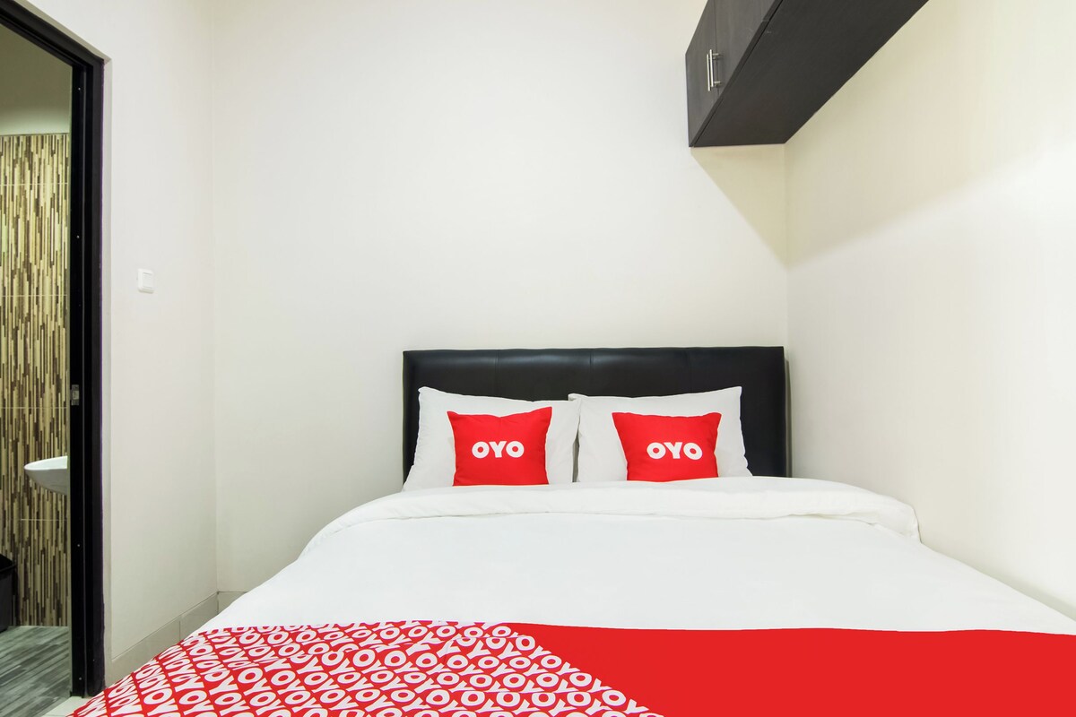 Una habitación de hotel con una cama, dos almohadas rojas con el texto "OYO", una colcha blanca y una manta estampada en rojo y blanco.