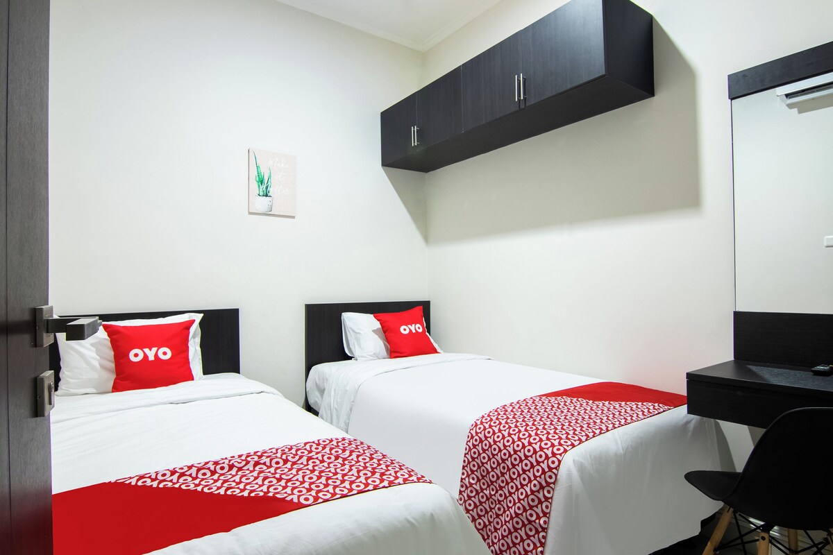 Habitación de hotel con dos camas, cada una con colcha de diseño rojo y blanco, cabecero negro y almohada roja con la palabra 'OYO'. Hay un escritorio con una silla negra, un armario negro sobre el escritorio y un cuadro en la pared.