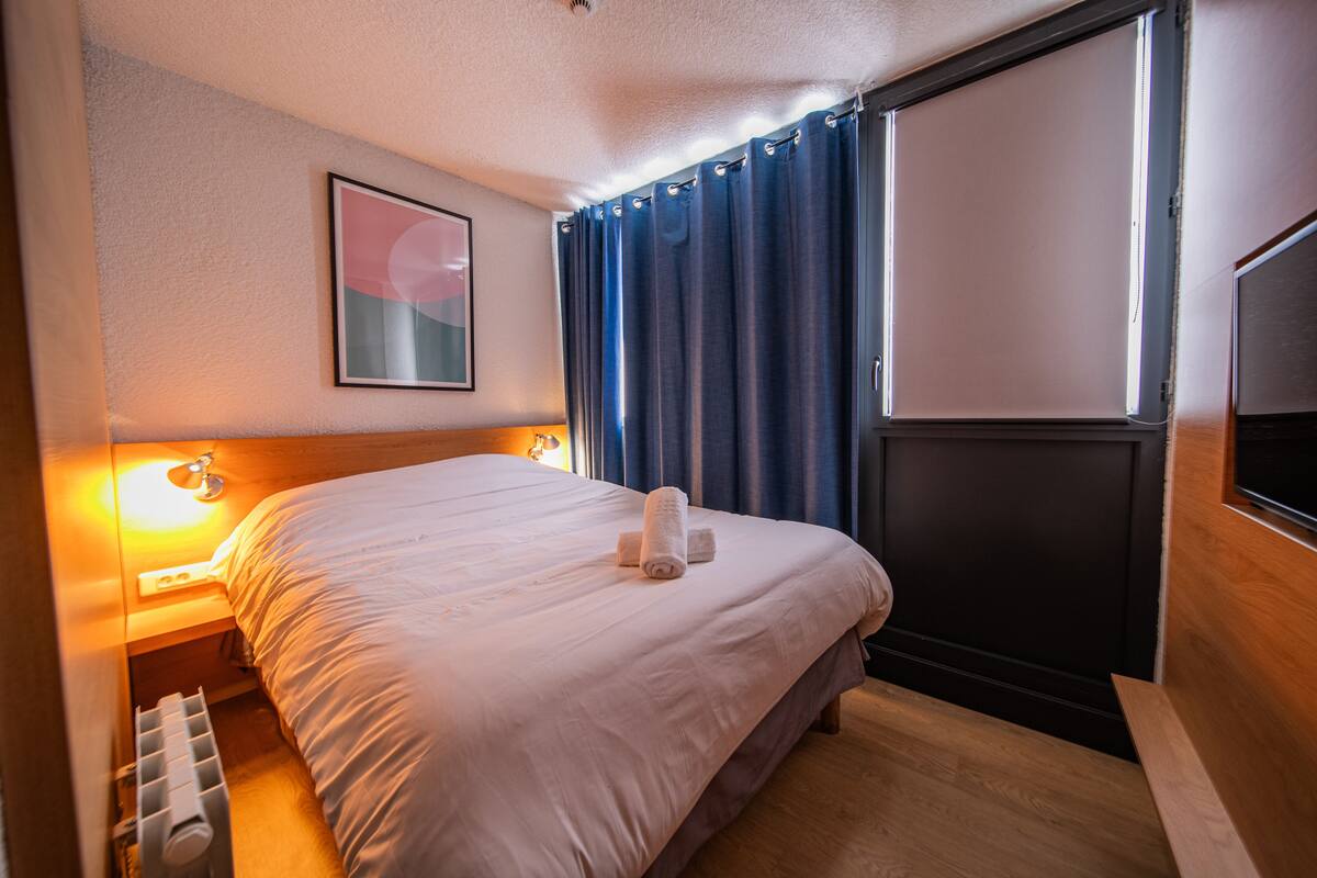Hotellihuone, jossa on sänky, yöpöytälamppu, ikkuna verhoilla ja seinälle asennettu televisio.
