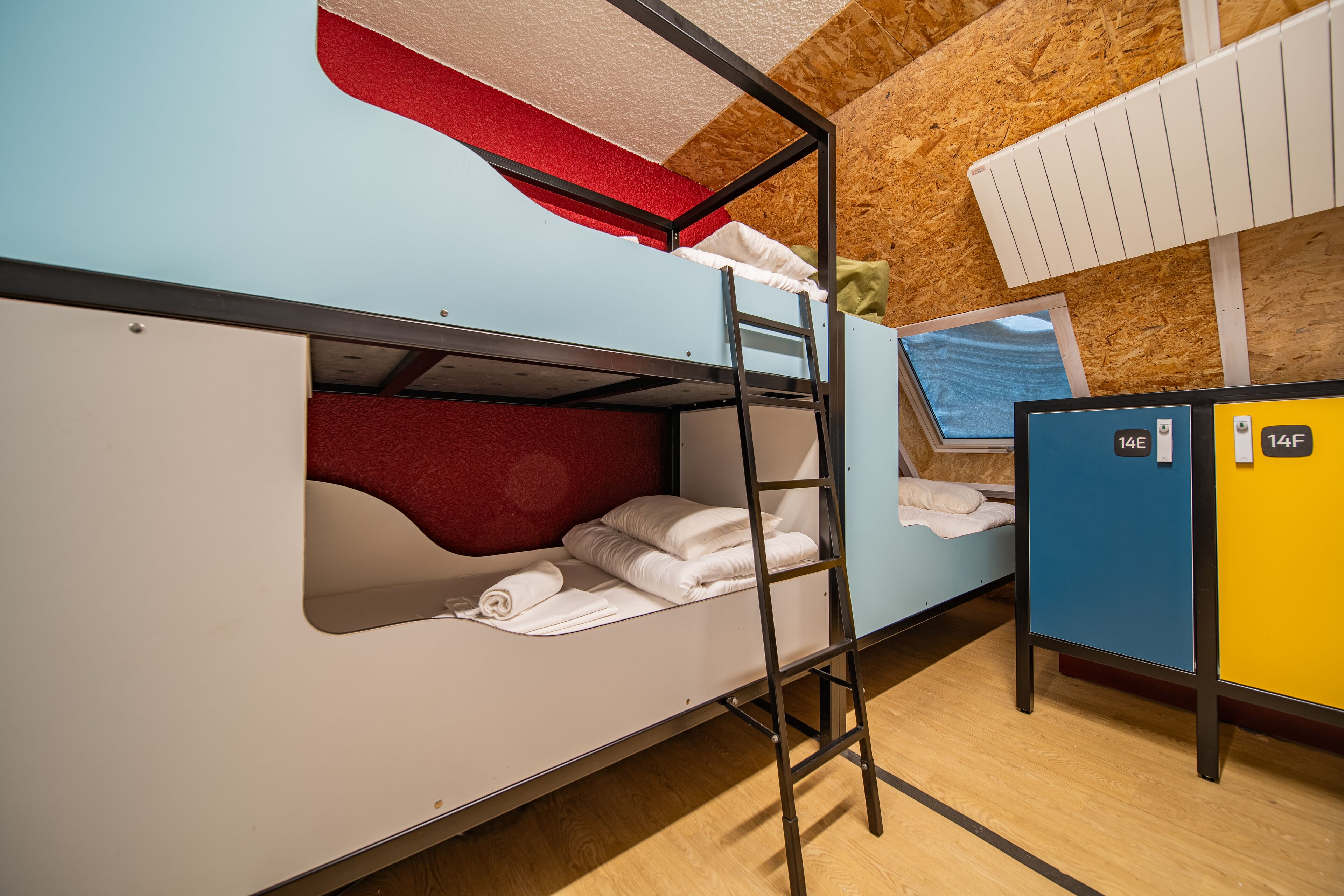 A bunk bed with a ladder, a red headboard, and a window. There are two lockers, one blue and one yellow, with numbers 14E and 14F.