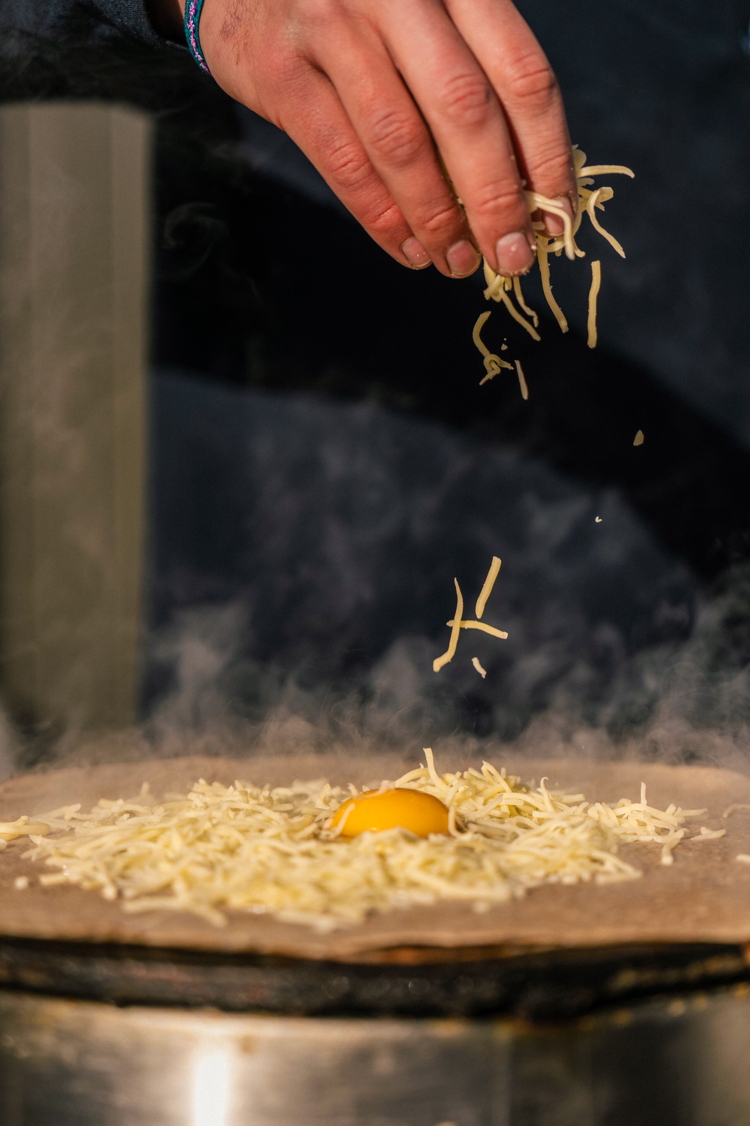 Une main saupoudre du fromage râpé sur une galette, qui est elle-même garnie d’un œuf.