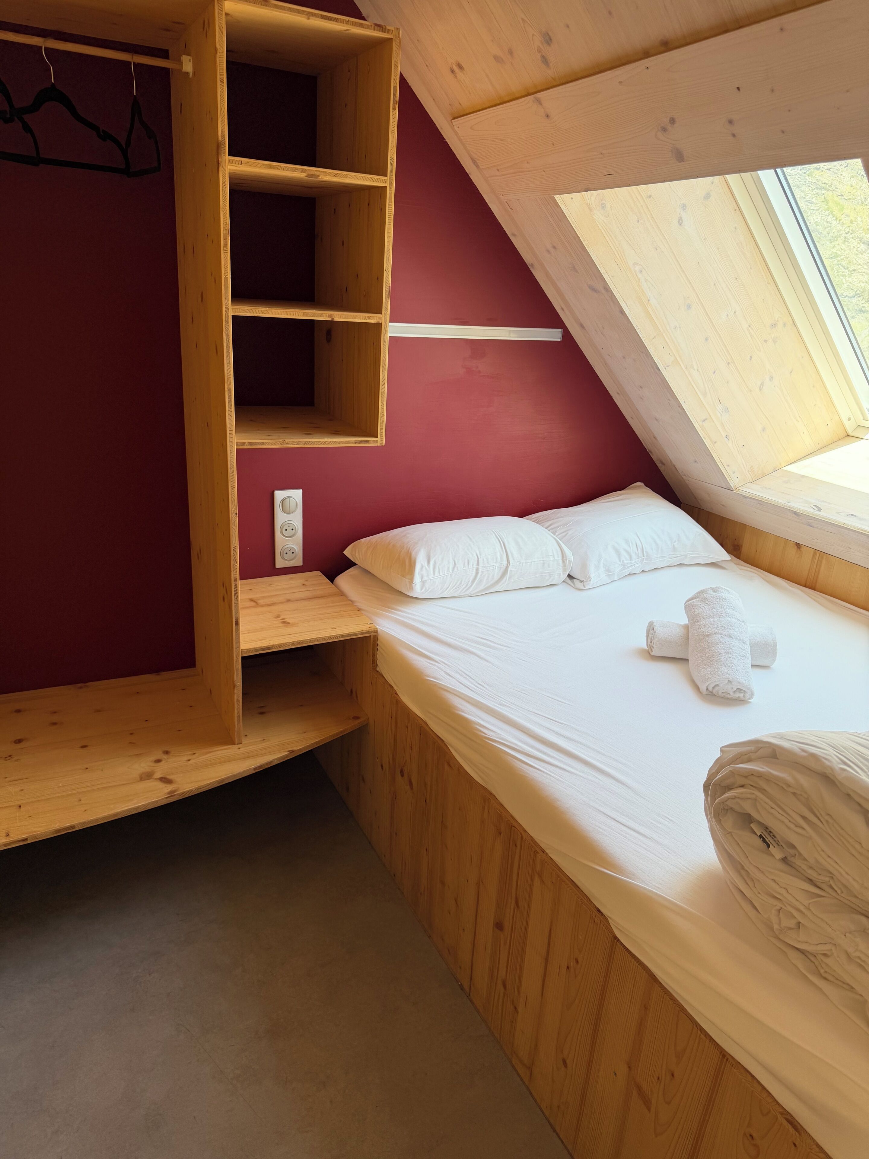A room with a sloped ceiling, a bed with white bedding, a wooden wardrobe, and a window providing natural light.