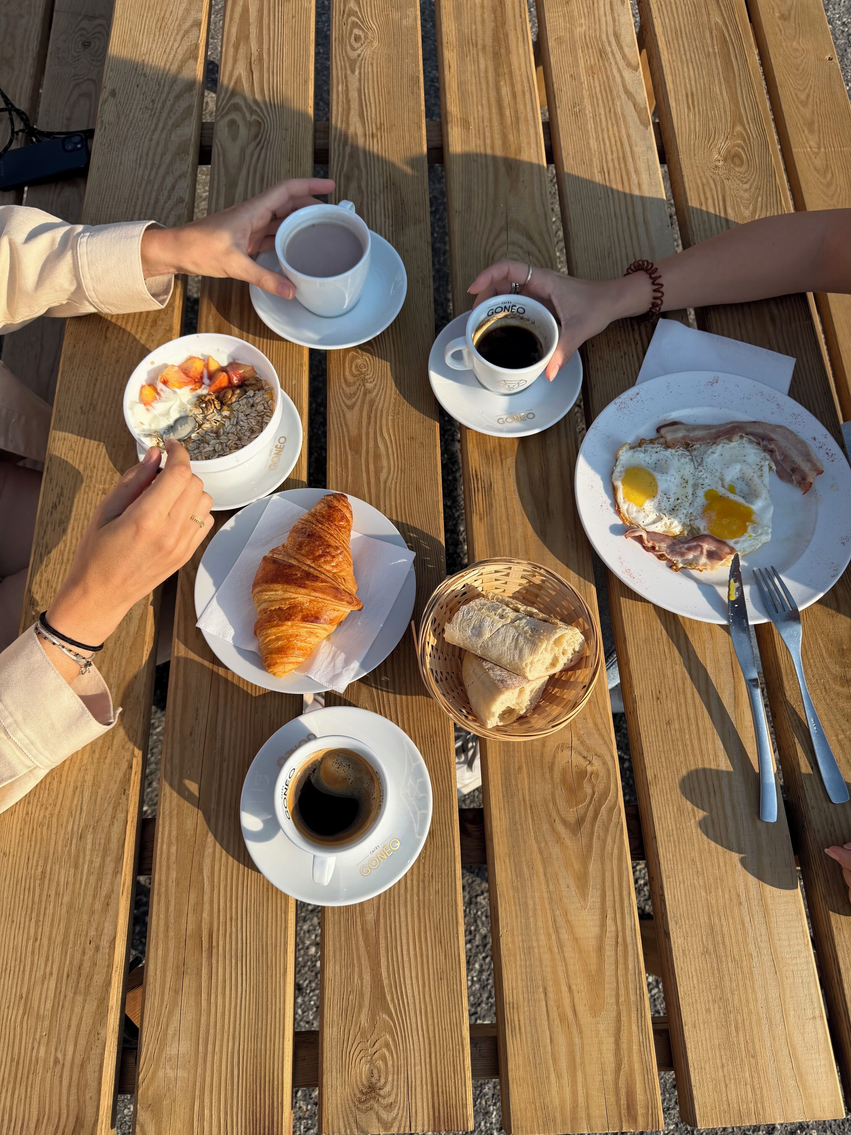 A wooden table with plates of food, cups of coffee, and a basket of bread.