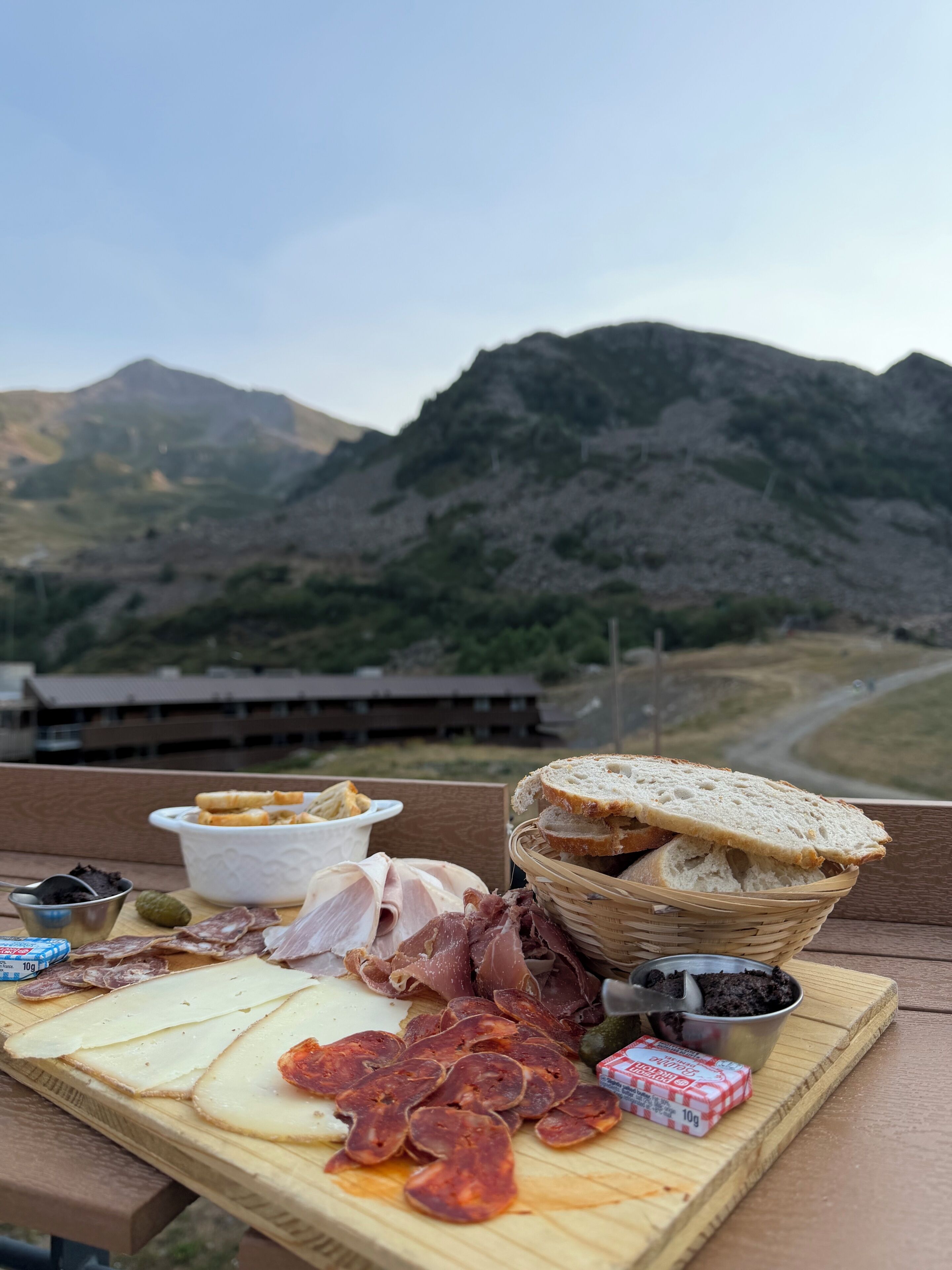 Un pique-nique avec différentes sortes de viandes, de fromages et de pain, disposés sur une table en bois, avec une vue sur les montagnes en arrière-plan.