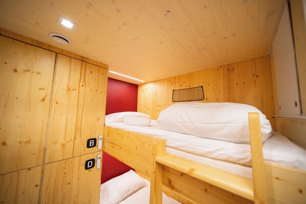 Una cama litera con estructura de madera y ropa de cama blanca, una pared roja y un techo con luz empotrada.
