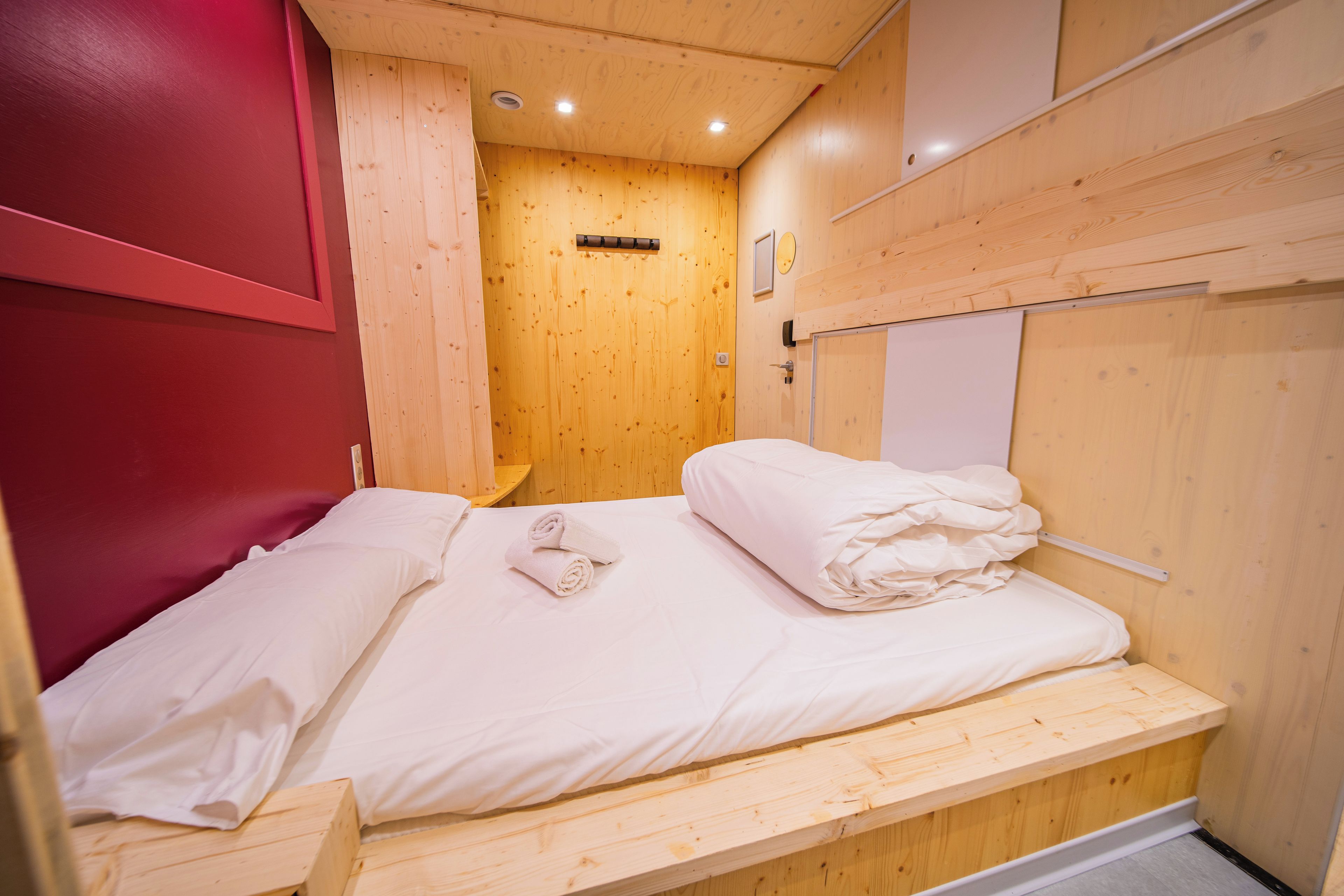 A small, compact room with a single bed, a wooden bench, and a red wall.