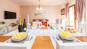 Un comedor con una mesa lista para cuatro personas, un sofá al fondo y un televisor montado en la pared.