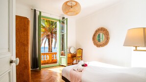 A bedroom with a bed, a wooden door, a mirror, and a view of palm trees and the sea through an open door.