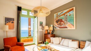 A room with a view of the sea, a wicker chair, a sofa, a table with a plant, and a painting of a boat.
