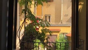 On aperçoit, à travers une fenêtre, un bâtiment à la façade jaune, aux volets rouges et dont la cheminée est bien visible.