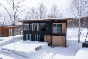 A modern, two-story cabin with large windows and a wooden deck, surrounded by snow. - Yuki Sugi at Intuition (Kutchan)