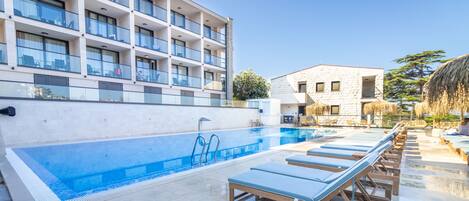 A swimming pool with lounge chairs, a building with balconies, and a clear sky.