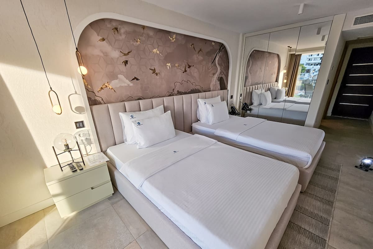 Ein Hotelzimmer mit zwei Betten, einem Nachttisch mit Lampe, einem Spiegel und Blick nach draußen.