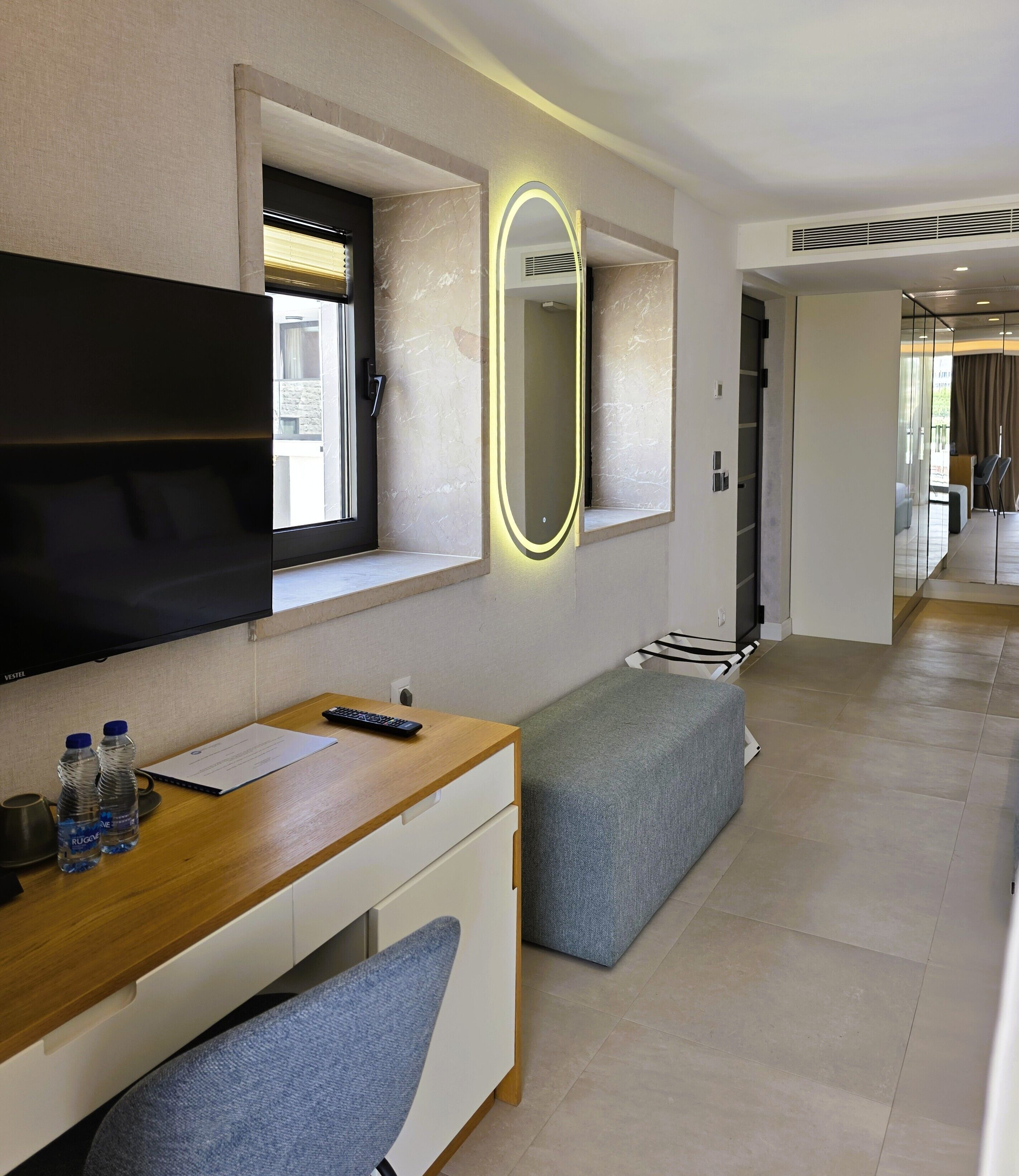Een moderne hotelkamer met een flatscreen televisie, een bureau met stoel, een bankje en een grote spiegel met verlichting.