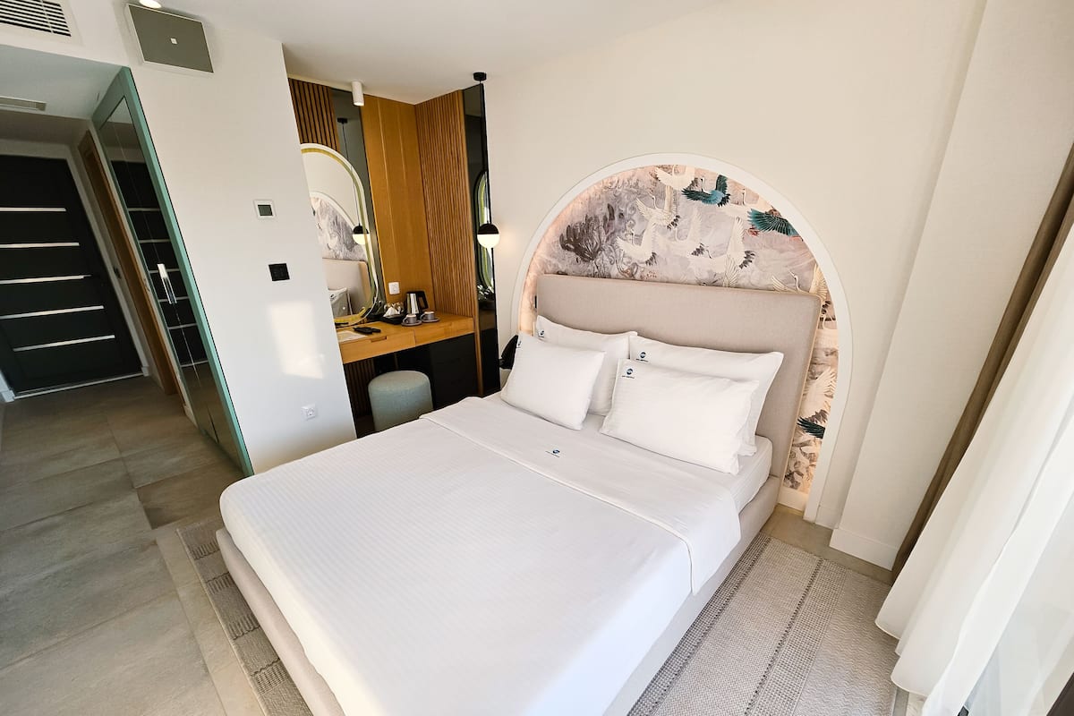 Ein modernes Hotelzimmer mit einem großen Bett, einem Schreibtisch mit Stuhl, einem Spiegel und einem kleinen Hocker.