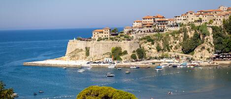 A coastal town with buildings on a cliff overlooking the sea, boats, and a beach area.