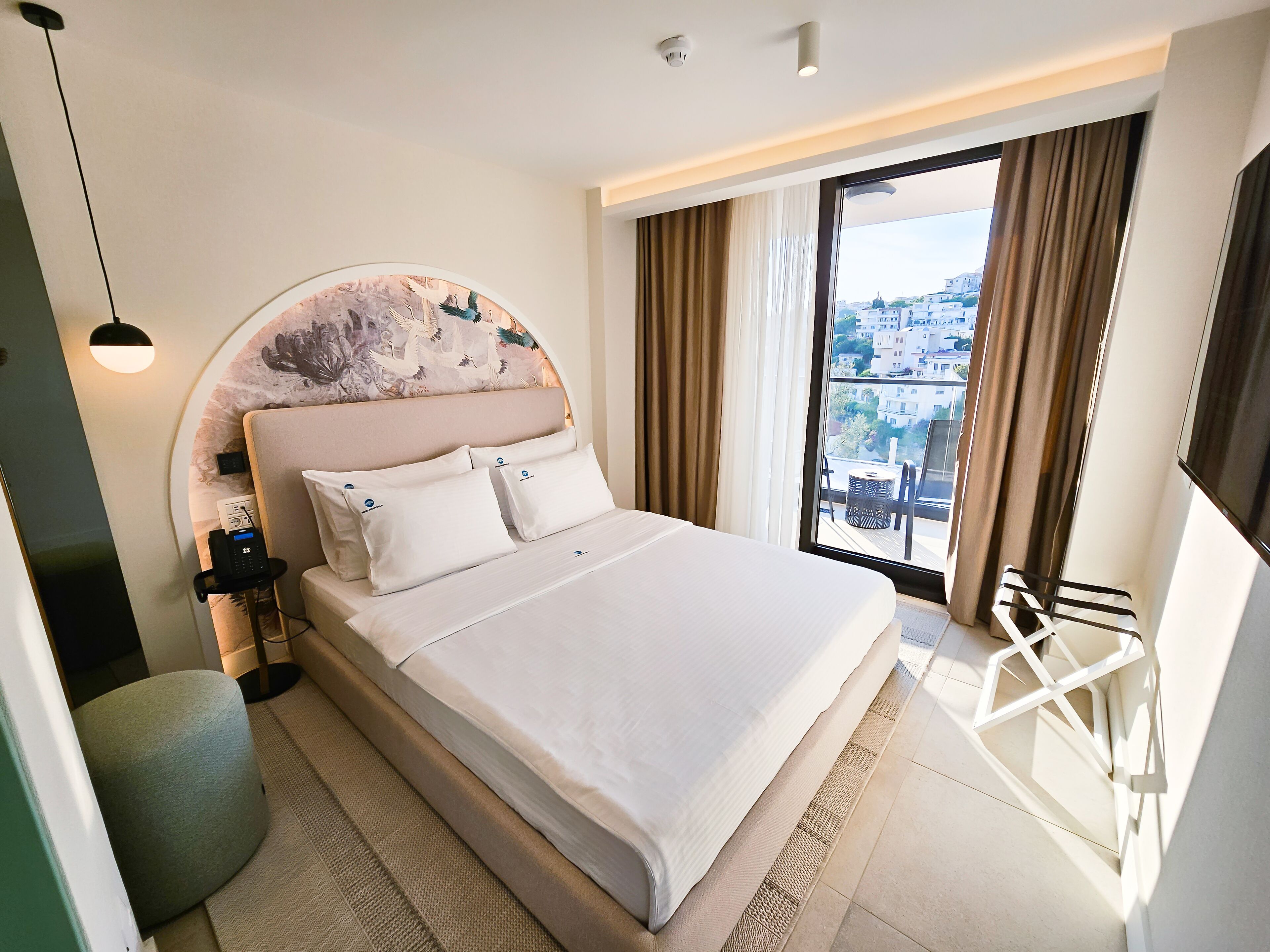 Une chambre d’hôtel moderne dotée d’un grand lit, d’une tête de lit ronde agrémentée d’un panneau décoratif, d’une table de chevet avec une lampe et offrant une vue sur la ville grâce à une grande fenêtre.