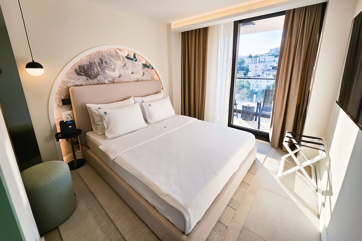 Ein modernes Hotelzimmer mit einem großen Bett, einem runden Kopfteil mit einer Zierleiste, einem Nachttisch mit einer Lampe und Blick auf die Stadt durch ein großes Fenster.