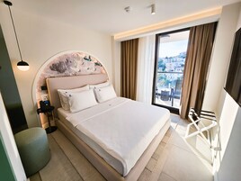 Una habitación de hotel moderna con una cama grande, cabecera redonda con panel decorativo, mesita de noche con lámpara y vistas a la ciudad a través de un ventanal amplio.