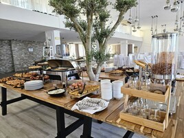 Montaje de bufé con diversos alimentos, una estación de autoservicio y un árbol decorativo en una habitación amplia con techo alto y decoración moderna.